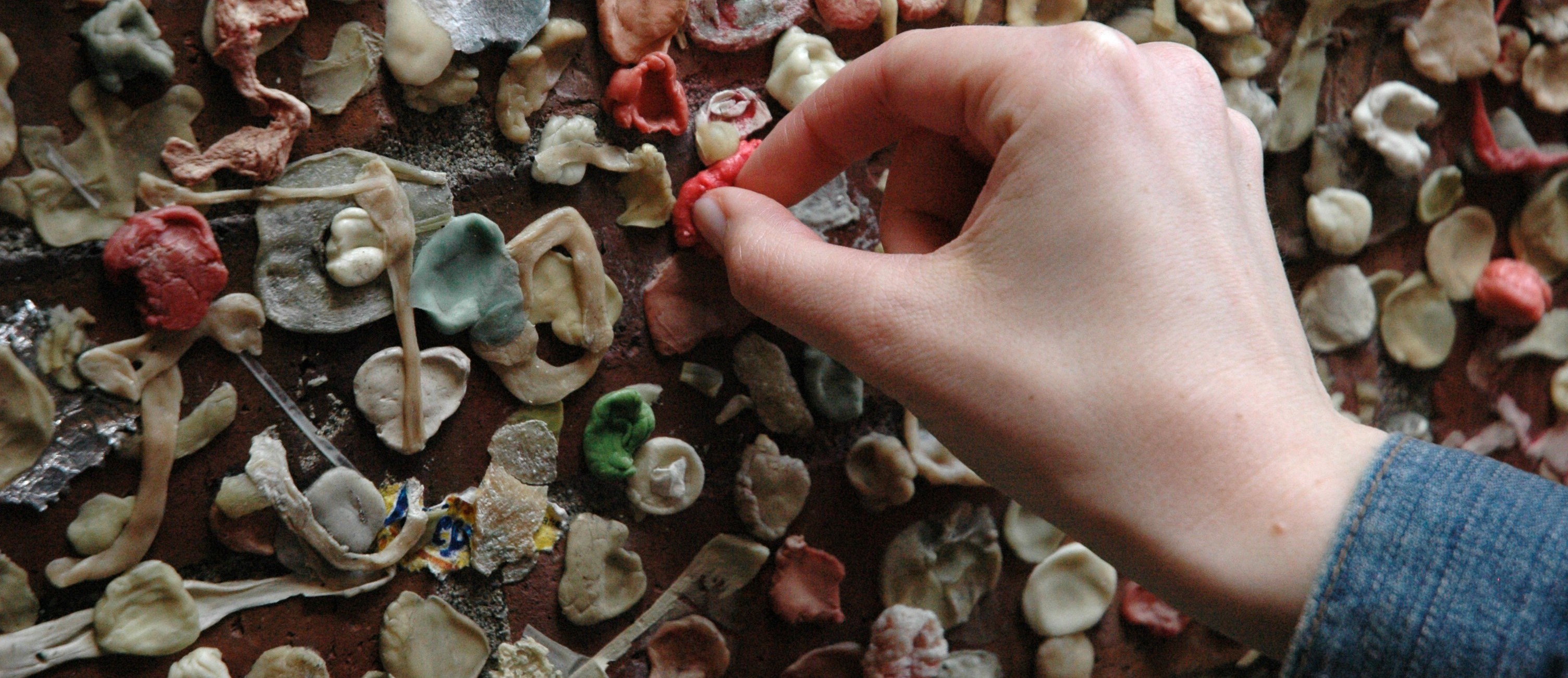 Contribute: hand adding freshly chewed piece of gum to a wall containing many differently colored pieces of chewed gum.